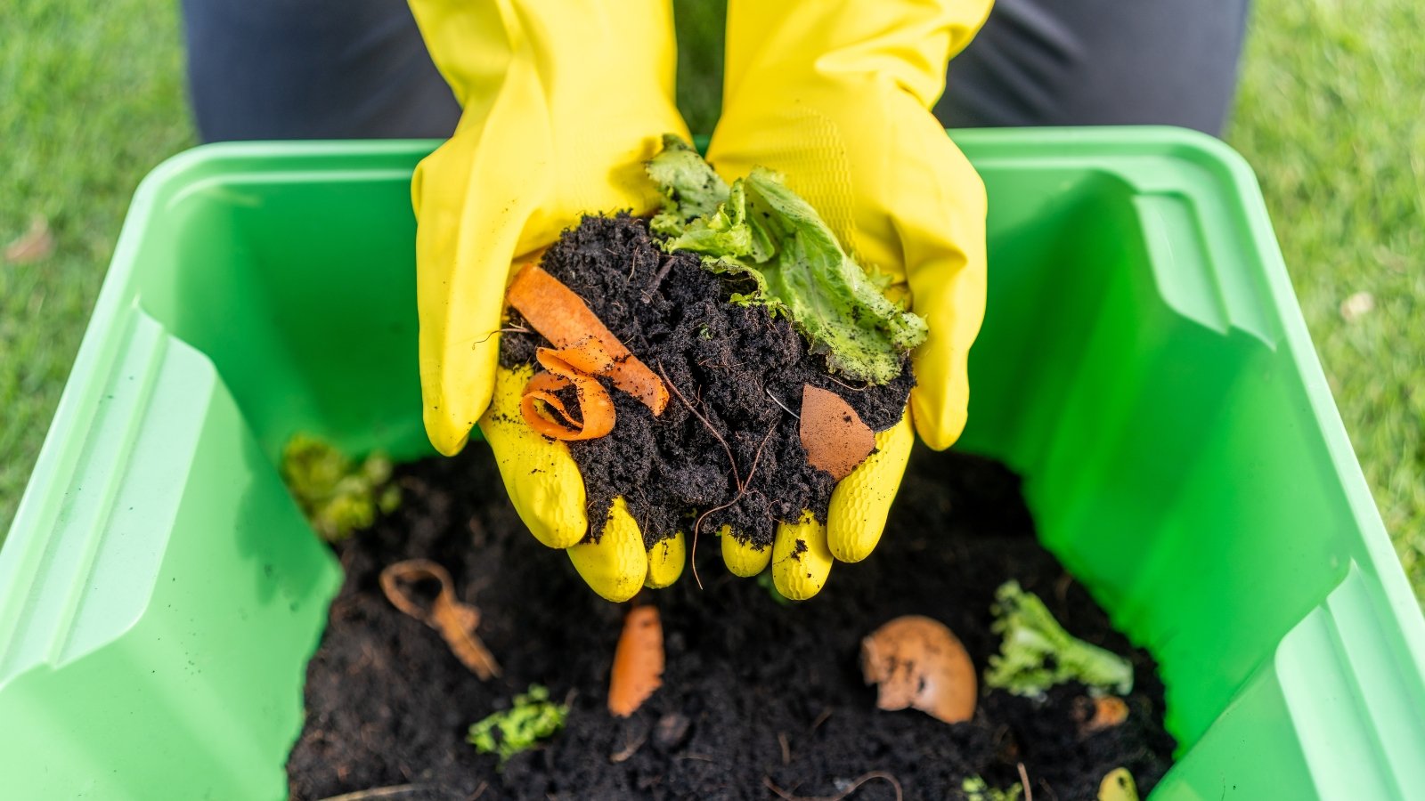 Bright yellow gloves hold a handful of fruit and vegetable scraps above a green bin filled with black soil and decomposing organic material.